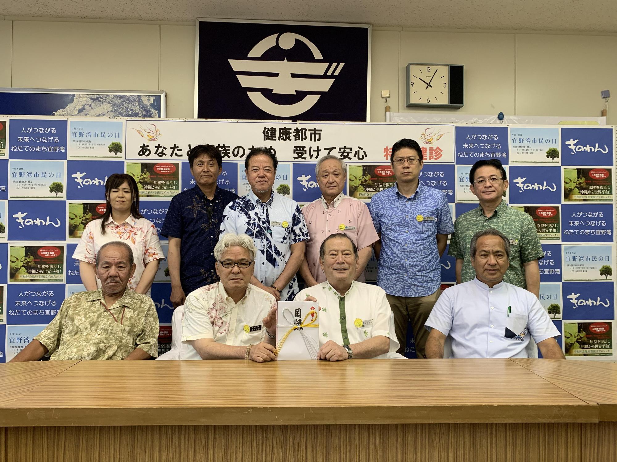 前列に4名の方々が座り、真ん中に座っている松川市長と1名の白髪の男性が目録を持っており、後方には6名の関係者の方々が笑顔で写っている集合写真