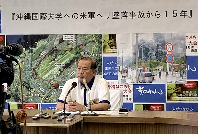 『沖縄国際大学への米軍ヘリ墜落事故から15年』と書かれた紙や、煙が立ち上っている写真が貼られたパネルを背景にテレビカメラの前で席に座り話をしている市長の写真
