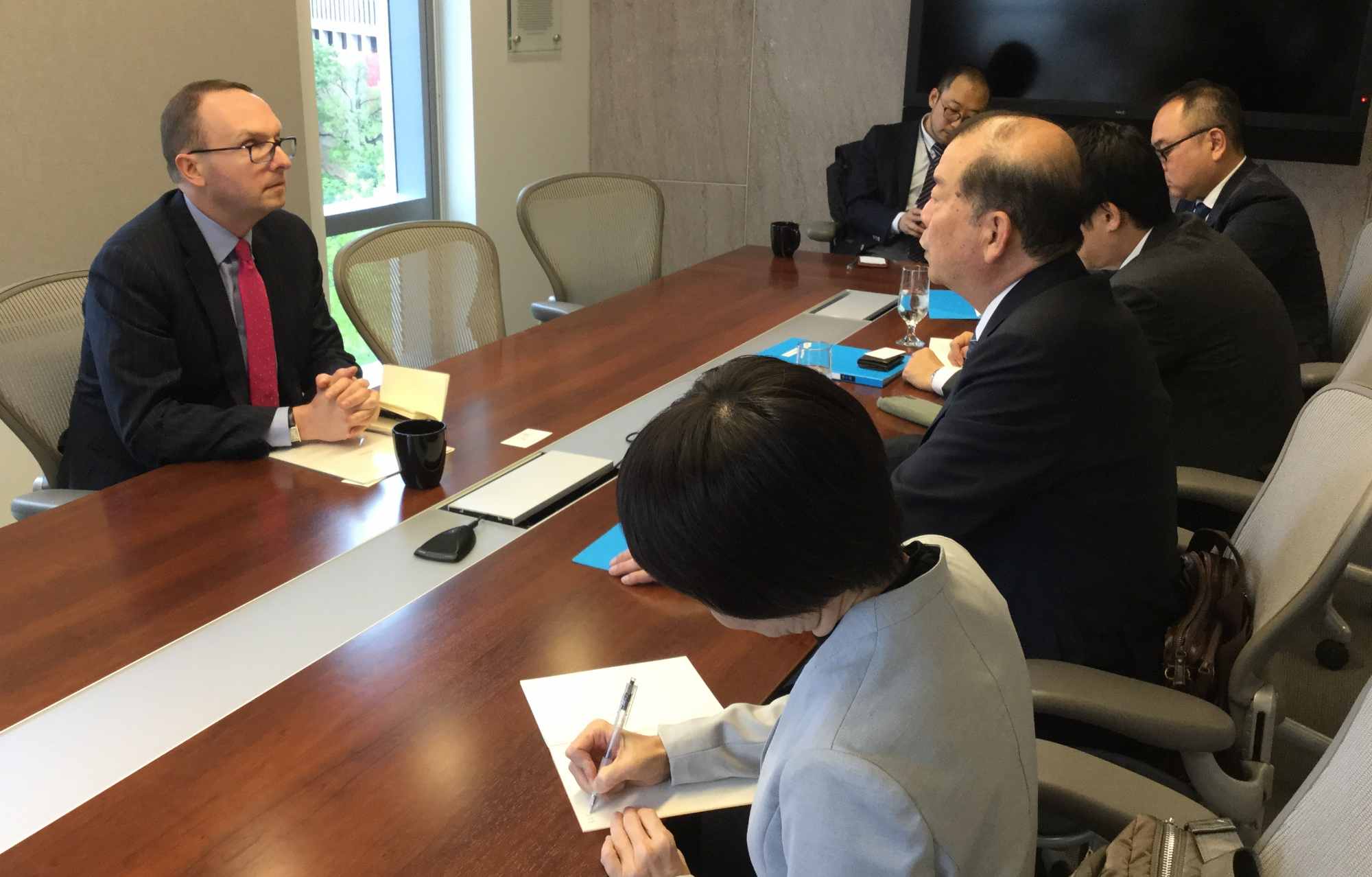 大きなテーブルを挟んで手前に日本人の女性1名と男性4名奥に1名、反対側に日本部副部長の男性が座って話しをしている写真