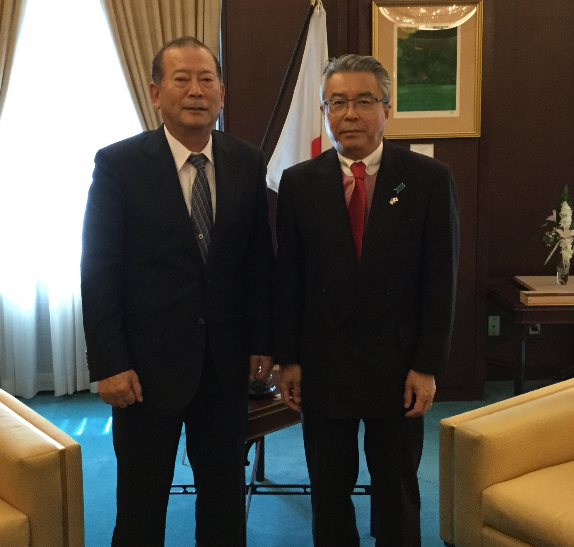 一人掛けのソファの間にスーツを着た男性2名が並んで立っている写真