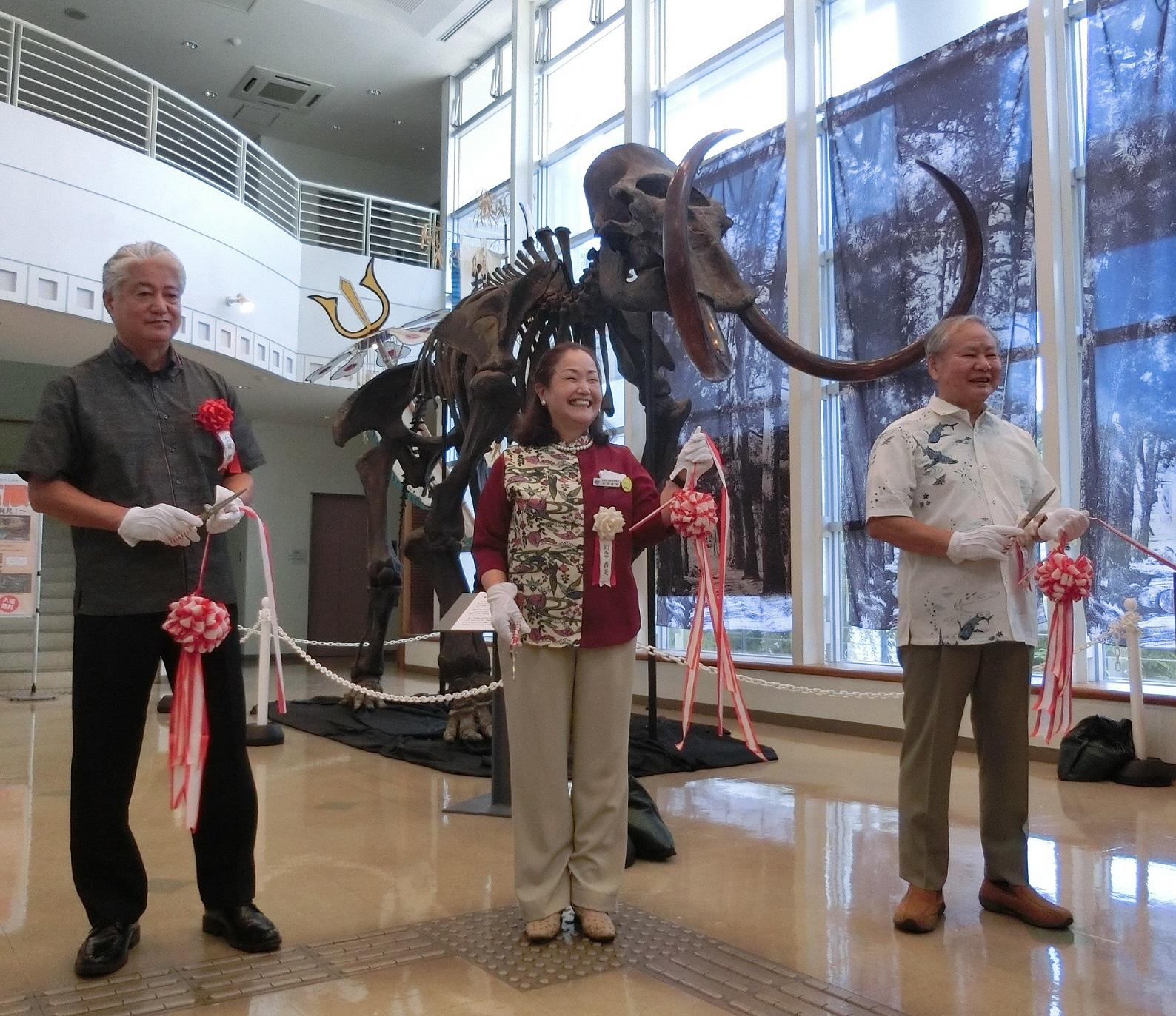 ロビーに展示された大きな象の化石の前で3名の来賓の方がテープカットをしている写真