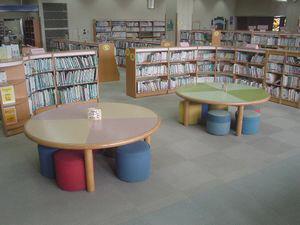 児童用の低めの本棚、丸テーブルと赤や青のかわいい椅子が置かれている写真