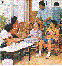 籐で造られた3人掛けの椅子の真ん中におばあちゃんと子供たちが座り、後ろに男性が立って話しをしている写真